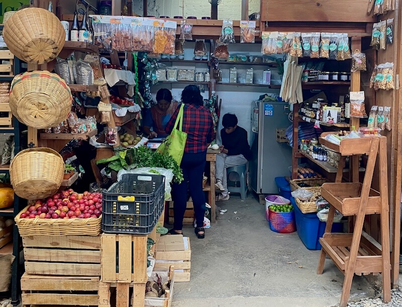 A person shopping at small shop that sells fruits, vegetables and other products. Wooden crates and woven baskets used for shopping and storage.