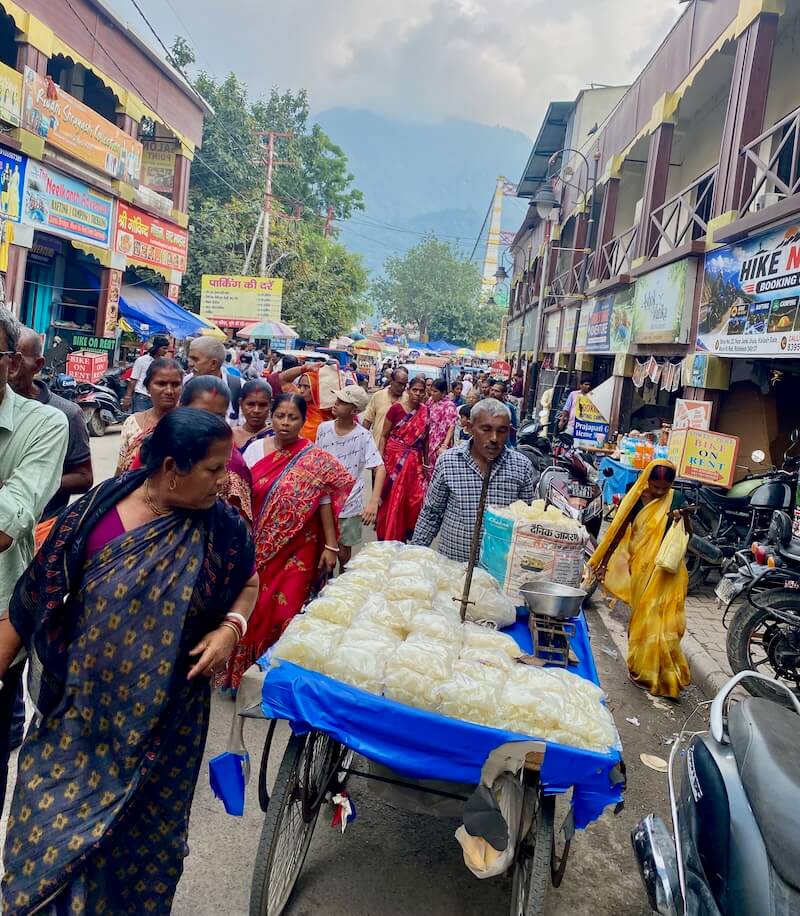 Busy Rishikesh street with many women in bright saree and a man pushing a large cart with snacks.
